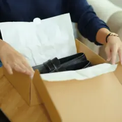 Kobieta przygotowuje zwrot towaru w sklepie stacjonarnym, pakując czarne mokasyny do kartonowego pudełka.