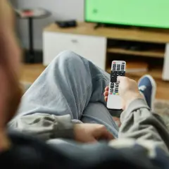 Osoba z pilotem w ręku, siedząca na kanapie przed zielonym ekranem telewizora. Zastanawia się, kto nie musi płacić abonamentu RTV.