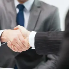 Dwóch mężczyzn w garniturach ściska sobie dłonie. Jeden z nich, w szarym garniturze, jest wyraźniejszy. To symboliczny gest zawarcia umowy, gdzie jeden z nich jest zleceniodawcą.