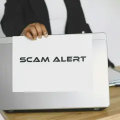 Ostrzeżenie przed oszustwem na Vinted. Ktoś trzyma kartkę z napisem "SCAM ALERT" nad laptopem.