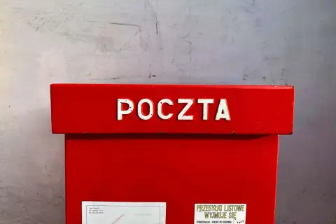 Czerwona skrzynka pocztowa z napisem "POCZTA". Informacja o godzinie odbioru przesyłek listowych: 14:00. Dowiedz się, ile kosztuje przelew na poczcie.