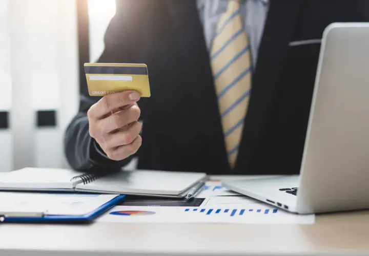 Biznesmen trzyma kartę kredytową, planując wykorzystać kredyt odnawialny do rozwoju firmy. Na biurku laptop i wykresy.