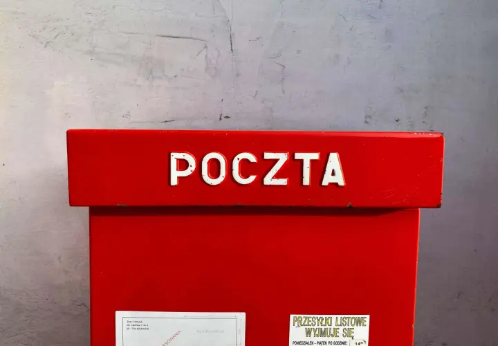 Czerwona skrzynka pocztowa z napisem "POCZTA". Informacja o godzinie odbioru przesyłek listowych: 14:00. Dowiedz się, ile kosztuje przelew na poczcie.