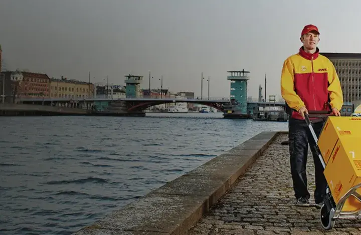 Kurier DHL z wózkiem pełnym paczek obok żółtego vana. Czy u kuriera DHL można płacić kartą? Tak, często jest to możliwe.