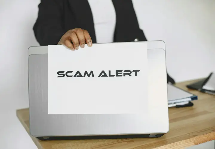 Ostrzeżenie przed oszustwem na Vinted. Ktoś trzyma kartkę z napisem "SCAM ALERT" nad laptopem.