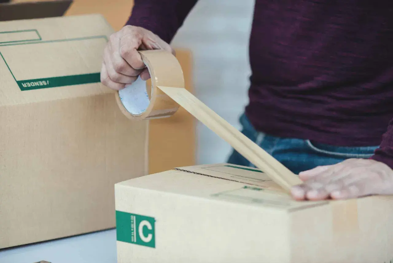Osoba zakleja taśmą kartonowe pudło. To część procesu przygotowania przesyłki, który może być związany z odprawą celną.