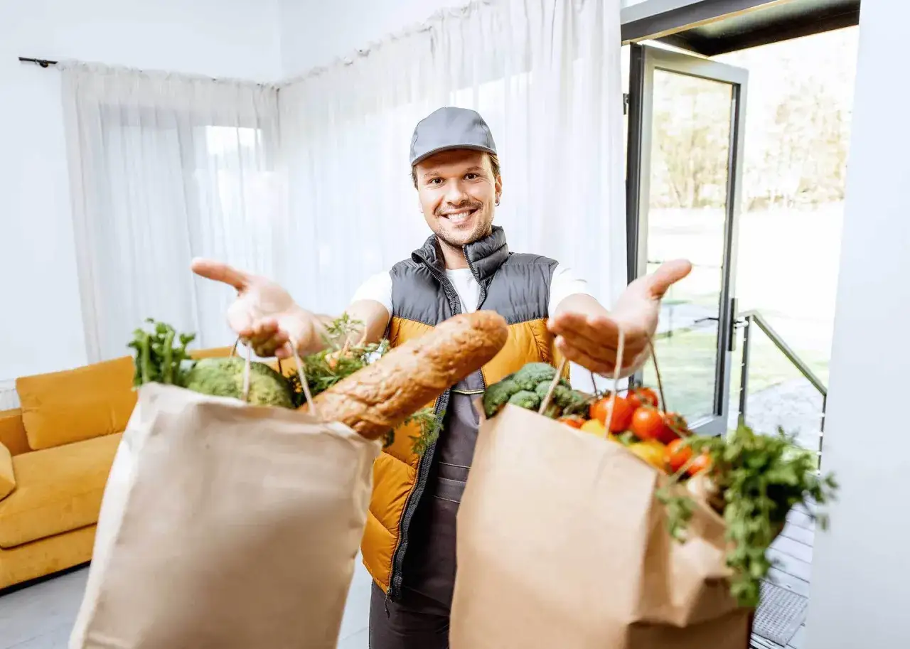 Uśmiechnięty kurier z dostawą b2c: świeże warzywa, pieczywo i owoce w papierowych torbach.
